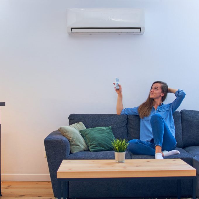 Young happy woman sitting on couch under air conditioner and adjusting comfort temperature with remote control at modern home